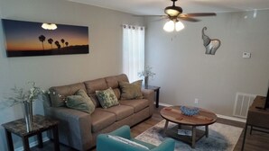 A living room with a brown sofa, teal armchair, and a coffee table. There is a ceiling fan, a painting of a sunset, and a small plant on a side table. - Quiet and Cozy Cottage -10 minutes from the beach! (Corpus Christi)