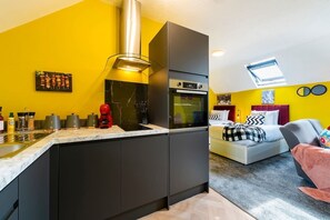 A compact kitchen with a built-in oven and a microwave, a dining area with a table and chairs, and a bed with a colorful headboard.