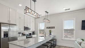 A modern kitchen with a stainless steel refrigerator, a microwave, and a coffee maker. There are bar stools along the island and a sign that reads "LAKE HOUSE". - Mallard Pointe 100 - Luxury condo- conveniently located! (Cullman)