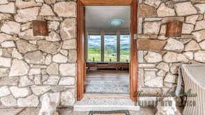 A stone-walled entrance with a wooden door, a view of greenery, and a wicker chair. - Tiger by the Tail Pets OK | Casita & Courtyard Oasis  w/ Heated Pool (Ruidoso Downs)