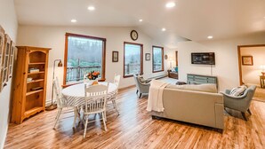 A spacious living room with a wooden floor, a dining table with chairs, a sofa, and a television mounted on the wall. - Cozy up in this private bungalow in the trees close to trailheads & shopping (Kodiak)