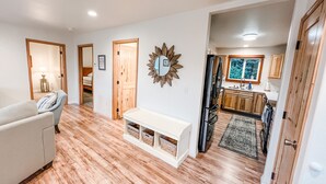 A spacious living area with hardwood flooring, a white sofa, a bench with storage, a kitchen with wooden cabinets, and a large window. - Cozy up in this private bungalow in the trees close to trailheads & shopping (Kodiak)