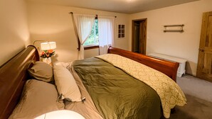 A bedroom with a bed, a bedside table with a lamp, a window with curtains, a wooden door, and a small shelf. - Cozy up in this private bungalow in the trees close to trailheads & shopping (Kodiak)