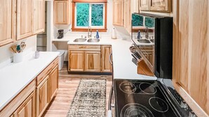 A kitchen with wooden cabinets, a stove, a sink, and a window with a view of trees. - Cozy up in this private bungalow in the trees close to trailheads & shopping (Kodiak)