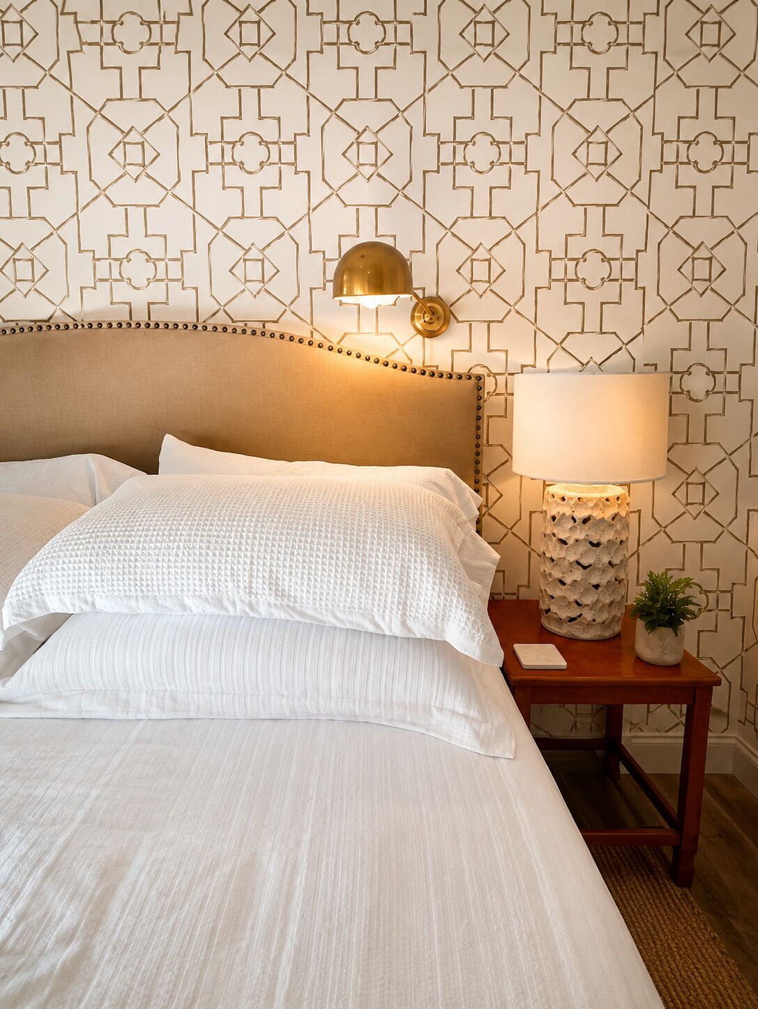 A neatly made bed with white linens, a headboard with a decorative trim, a bedside lamp, and a small table with a plant.