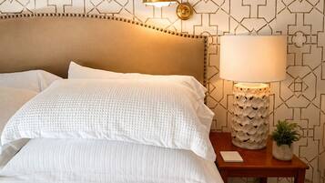 A neatly made bed with white linens, a headboard with a decorative trim, a bedside lamp, and a small table with a plant.