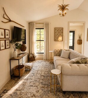 A living room with a sofa, a television, a chandelier, and a large window with curtains. - secluded cottage- firepit, outdoor shower, hammock (Readyville)