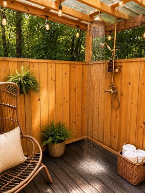 Una terraza de madera con ducha integrada, una silla de mimbre y una cesta de toallas.