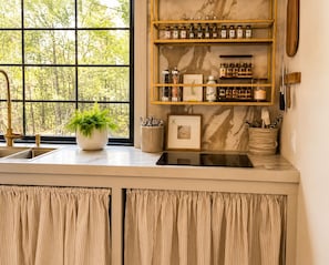 A kitchen with a marble backsplash, wooden shelves with jars, a potted plant, and a window with a view of trees.