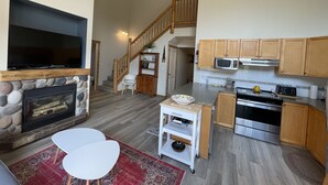 A spacious living area with a fireplace, a TV, and a dining table. A kitchen with wooden cabinets and modern appliances is visible in the background. - 5 Min. Drive to the Resort and Fernie (Fernie)