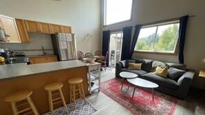 A modern kitchen with wooden cabinets and a stainless steel refrigerator, a cozy living area with a grey sofa and a red rug, and a large window with curtains. - 5 Min. Drive to the Resort and Fernie (Fernie)