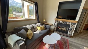 A living room with a sofa, a flat-screen TV, a stone fireplace, and a view of mountains through a window. - 5 Min. Drive to the Resort and Fernie (Fernie)