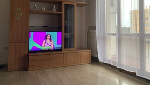 Una sala de estar con un mueble de madera para entretenimiento, un televisor mostrando un programa y un ventanal con cortinas translúcidas.