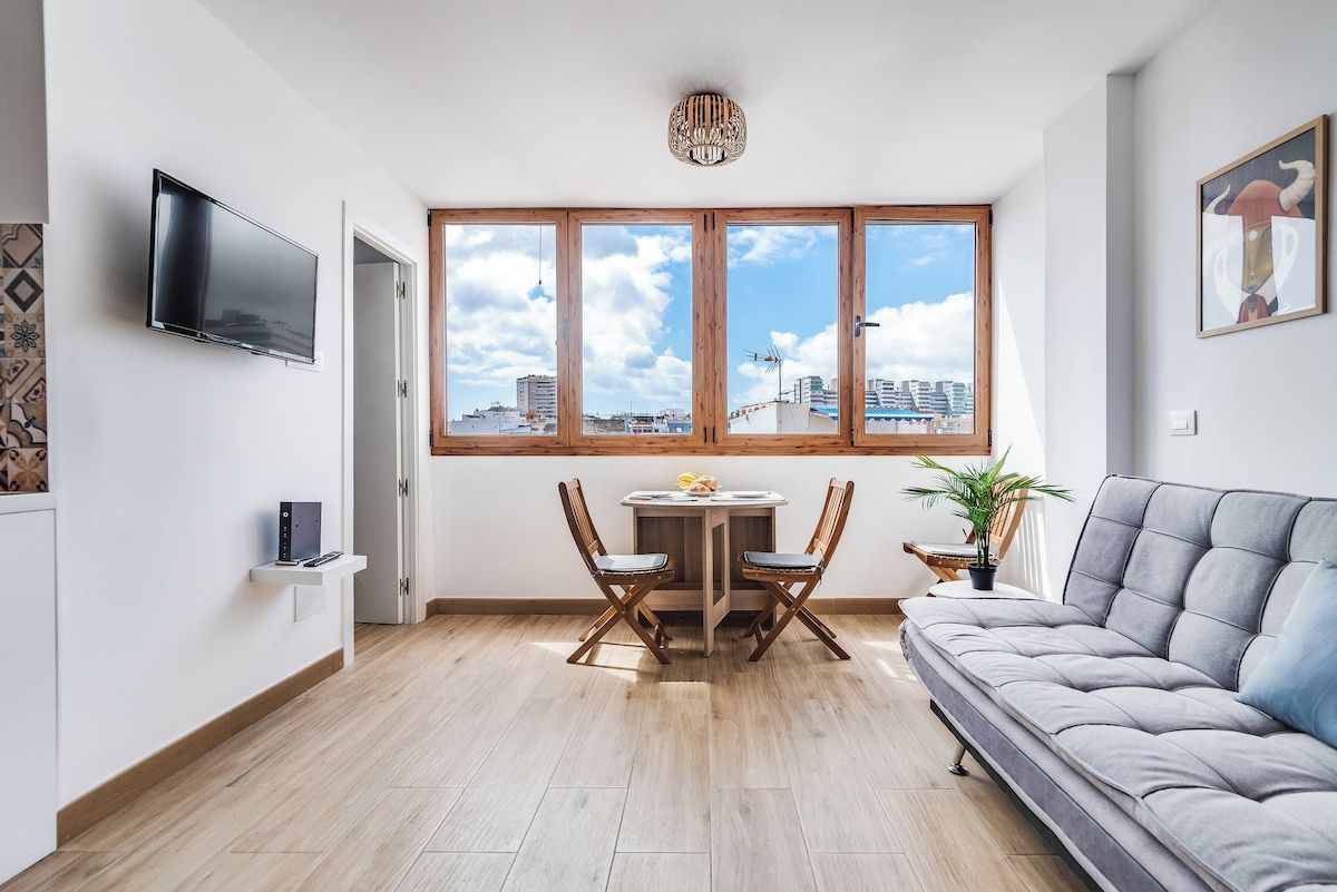 A modern living room with a grey sofa, a dining table with chairs, a TV mounted on the wall, and a large window with a city view.