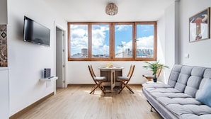 Ett modernt vardagsrum med en grå soffa, ett matbord i trä med två stolar, en TV som är monterad på väggen och stora fönster med utsikt över staden.