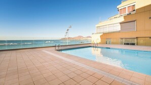En pool med utsikt över havet och kullarna, ett kaklat poolområde och en byggnad med stora fönster och en balkong.