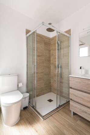 Une salle de bain moderne avec une douche à parois de verre, des toilettes blanches, une poubelle et un meuble-lavabo en bois avec un miroir.