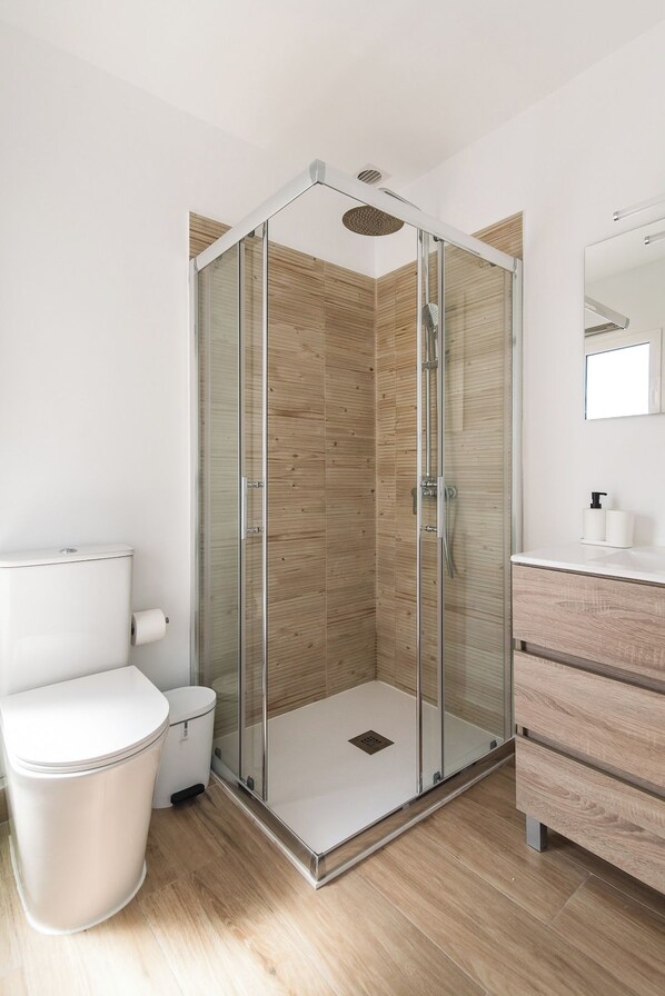 Un baño moderno con ducha envuelta en vidrio, inodoro blanco, un cesto de basura y tocador de madera con espejo.