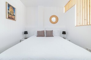 Une chambre à coucher avec un lit, deux tables de chevet avec des lampes, un miroir fixé au mur et un tableau encadré.