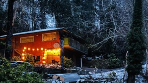 A cabin with a porch lit by string lights, surrounded by trees and snow.