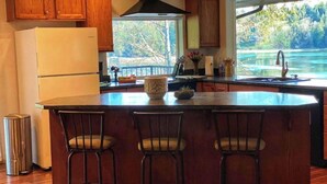 A kitchen with wooden cabinets, a large island with bar stools, a refrigerator, and a view of a lake through the window.