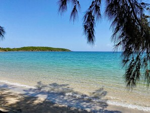 Une plage à l’eau turquoise et limpide, un rivage sablonneux et, en arrière-plan, une colline verdoyante.
