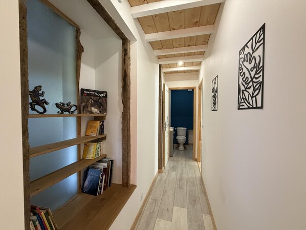 A narrow hallway with wooden beams, a bookshelf, and a blue door leading to a bathroom. - Gite DU Chene - Ledeuix (LEDEUIX)