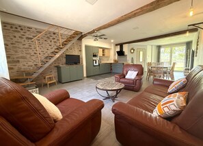 A spacious living area with leather sofas, a coffee table, a staircase with a wooden railing, a kitchen area, and a dining area with wooden furniture. - Gite DU Chene - Ledeuix (LEDEUIX)