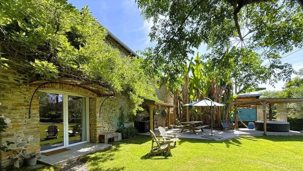 A stone house with a glass door, a patio with chairs, and a covered area with a table and umbrella. - Gite DU Chene - Ledeuix (LEDEUIX)