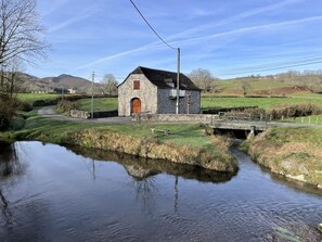 Un bâtiment en pierre avec une porte en bois, un petit pont enjambant une rivière paisible et un paysage rural avec des collines en arrière-plan.