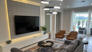 Una sala de estar moderna con un televisor de pantalla plana grande montado en una pared blanca, un sofá gris, dos sillones, una mesa de centro con objetos decorativos y una alfombra estampada.