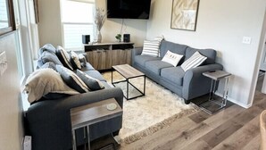 A modern living room with a grey sofa, a coffee table, and a TV mounted on the wall. - Gorgeous Beach House w/Pop Up Bar & Community Pool (Rockport)