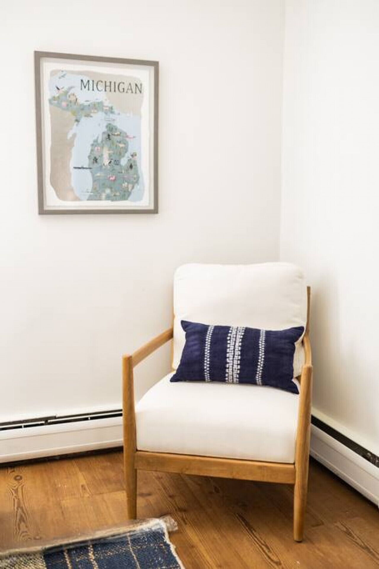 Un rincón de una habitación con un mapa enmarcado de Michigan, un sillón blanco con un cojín azul y un piso de madera.