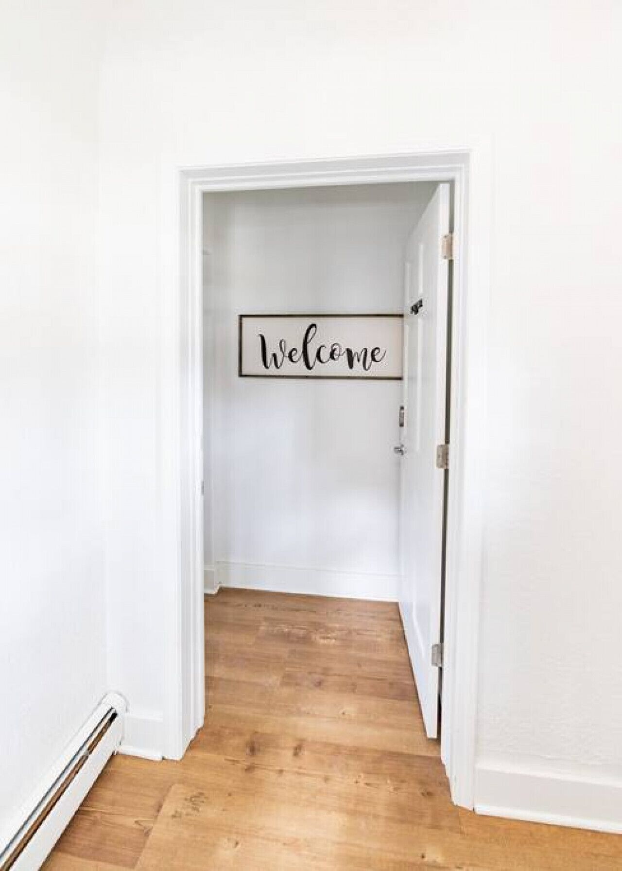 Een gang met een 'Welcome' bord aan de muur, houten vloer en witte wanden.