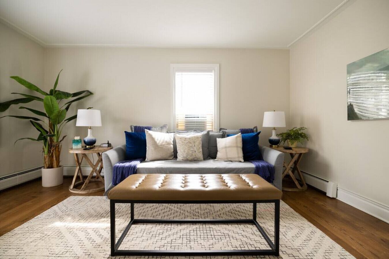 A living room with a sofa set, two side tables with lamps, a large potted plant, and a framed artwork on the wall.