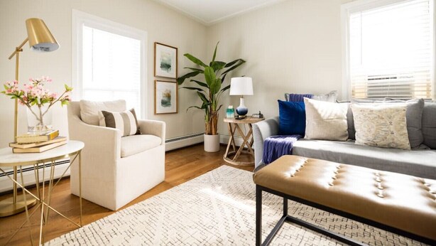A living room with a sofa, armchair, coffee table, and a lamp. There is a plant and a window with blinds. - Parsonage Suite - Millpond Inn (Clarkston)