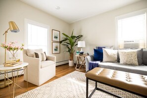 A living room with a sofa, armchair, coffee table, and a lamp. There is a plant and a window with blinds. - Parsonage Suite - Millpond Inn (Clarkston)