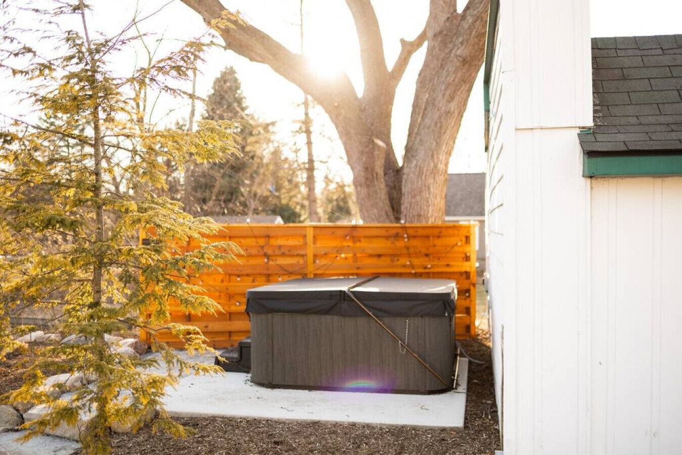 A backyard with a hot tub, a wooden fence, and a tree.