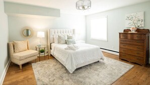 Een slaapkamer met een bed, een stoel, een dressoir, een spiegel en een raam.