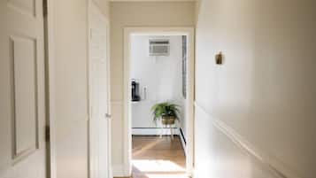 Un couloir aux murs blancs et au plancher en bois mène à une pièce dotée d’un ventilateur de plafond et d’une plante en pot.