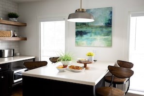 A modern kitchen with a central island, wooden chairs, a hanging light fixture, and a painting on the wall. - Parsonage and Main Street Suite - Millpond Inn (Clarkston)