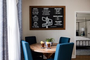 Un espace repas avec une table ronde, des chaises bleues et un tableau affichant le menu, qui propose des options pour le petit-déjeuner, le déjeuner, le dîner et des plats de cuisine de rue.
