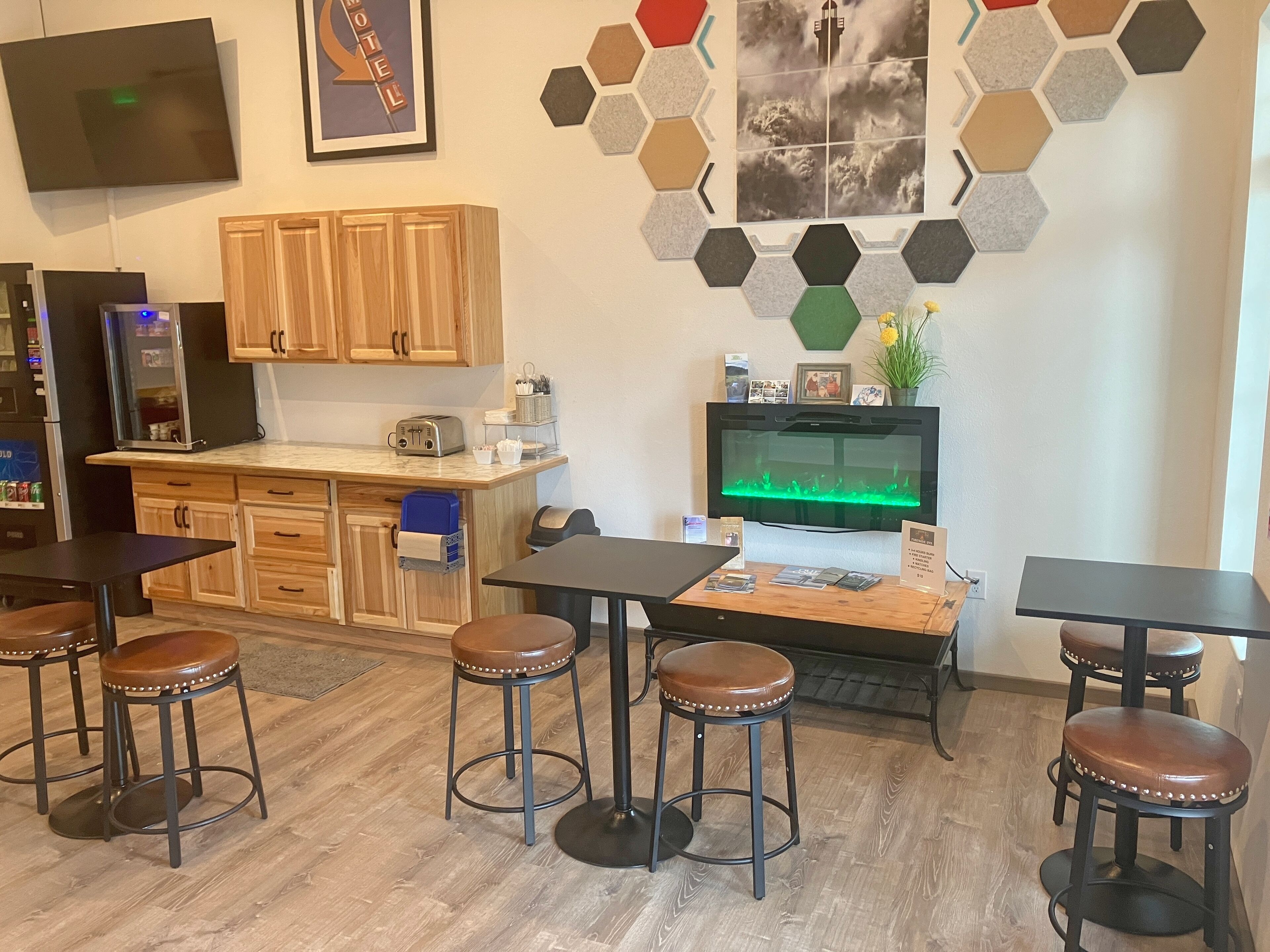 A modern kitchen with wooden cabinets, a built-in oven, and a coffee machine. There are bar stools and tables, and a large flat-screen TV mounted on the wall.