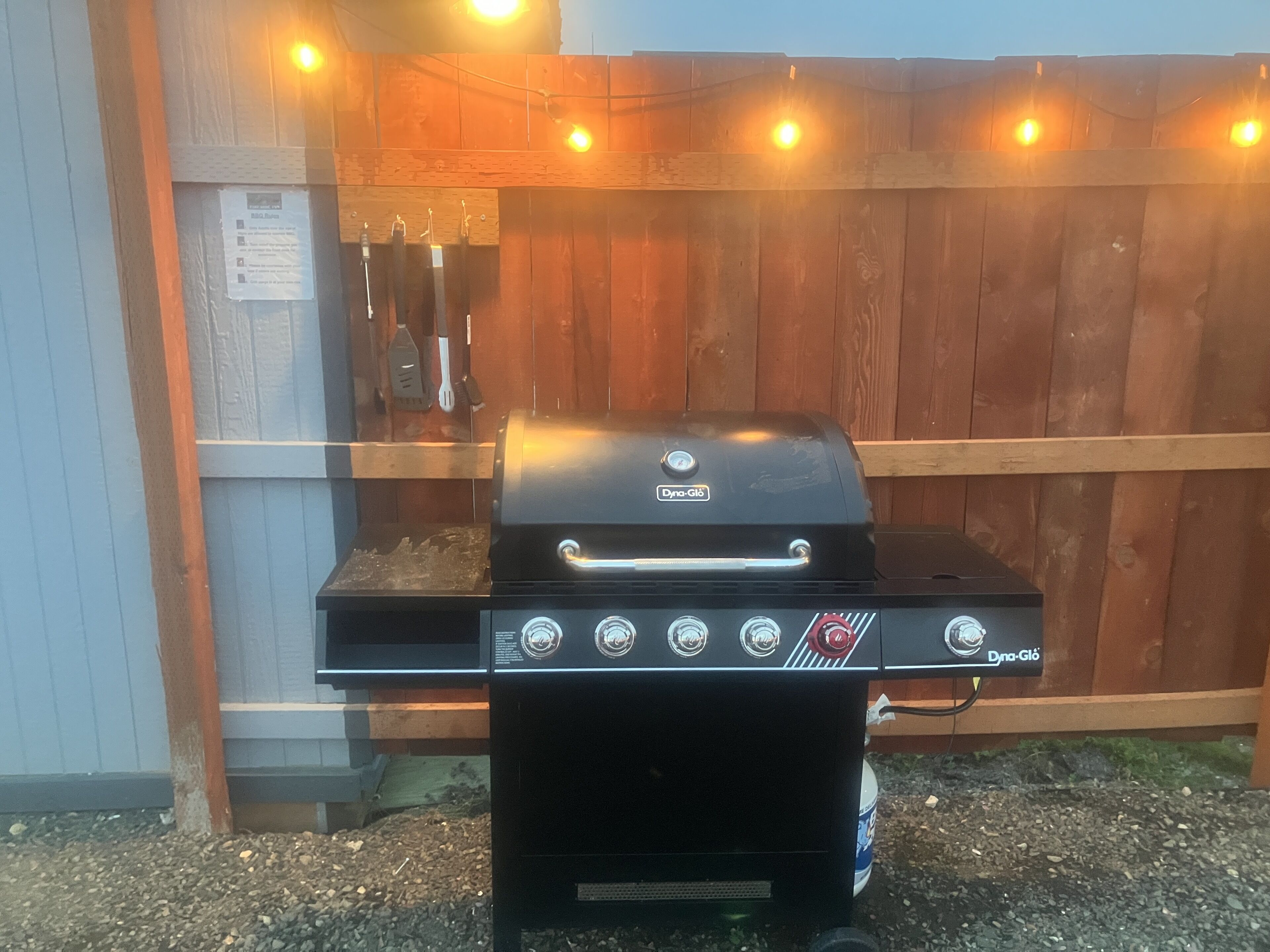 Una parrilla exterior negra con cuatro perillas de control y una pantalla digital, ubicada frente a una valla de madera con luces colgantes.