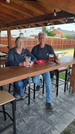 Two people seated at an outdoor wooden table with a red basket and drinks, under a wooden pergola with hanging lights.