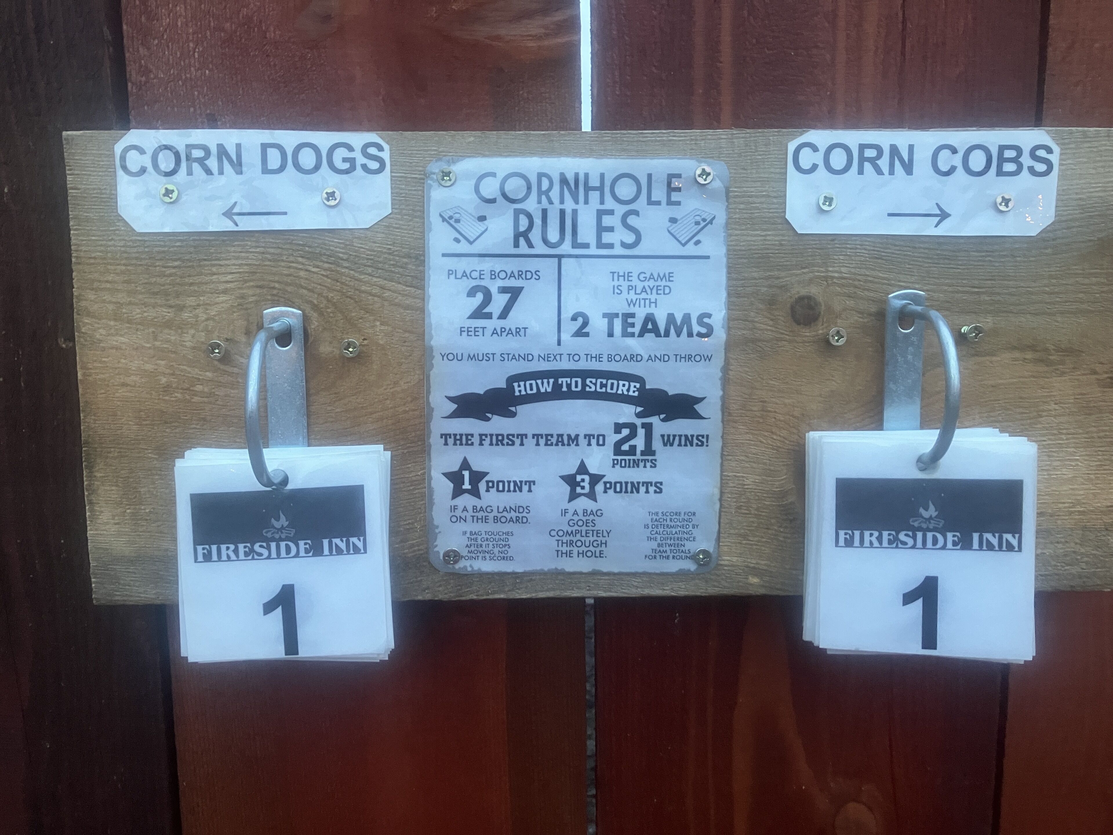 Una tabla de madera con letreros que dicen 'Corn Dogs', 'Reglas de Cornhole' y 'Mazorcas de Maíz', cada uno con flechas direccionales y el número '1'.