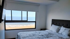 Une chambre avec une grande fenêtre donnant sur une plage, un lit recouvert d’une couvre-lit à motifs et une tête de lit sombre.
