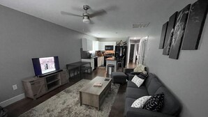 Ein modernes Wohnzimmer mit einem grauen Sofa, einem Couchtisch und einem Fernseher auf einem Holzboden. Im Hintergrund befindet sich eine Küche mit weißen Schränken und einem Essbereich.