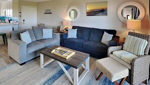 Una sala de estar con un sofá gris, un sofá azul, un sillón de mimbre y una mesa de centro. Hay un cuadro en la pared y un espejo redondo sobre la mesa de centro.