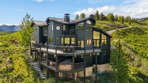 Et moderne hus med to etager, en terrasse, omgivet af grønt og bjerge.